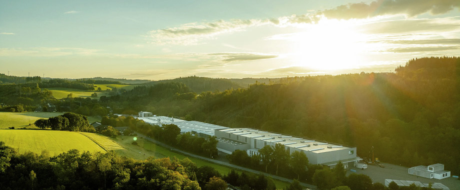 Industriegebäude in grüner Hügellandschaft bei Sonnenuntergang mit Wald, Feldern und weitläufigem Firmengelände im Vordergrund