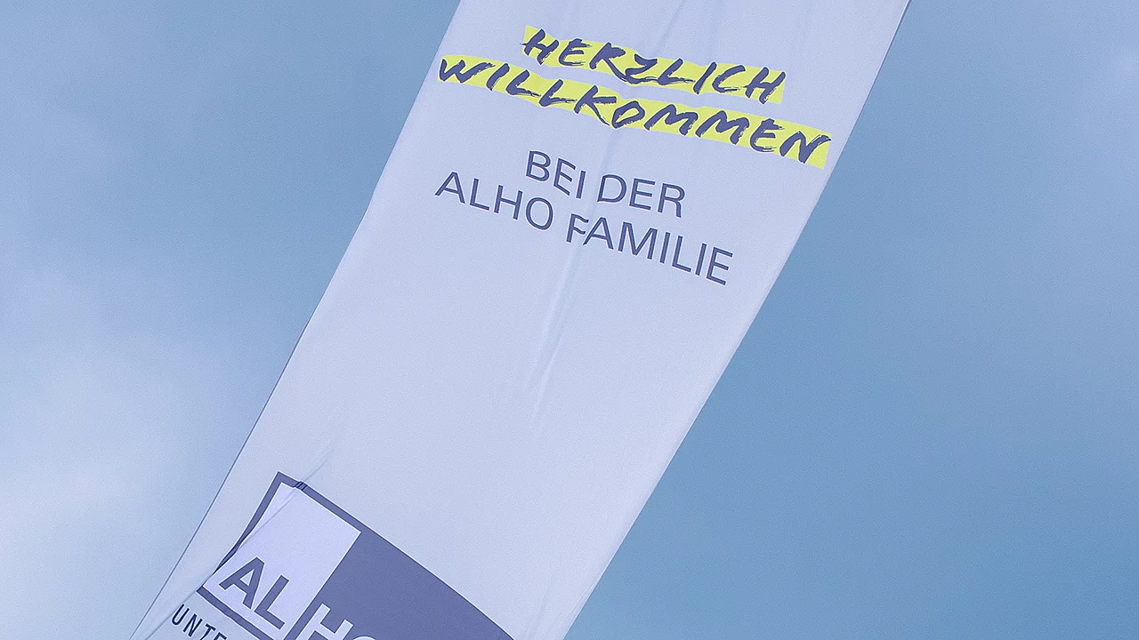 Fahne mit der Aufschrift „Herzlich willkommen bei der ALHO Familie“ vor blauem Himmel.