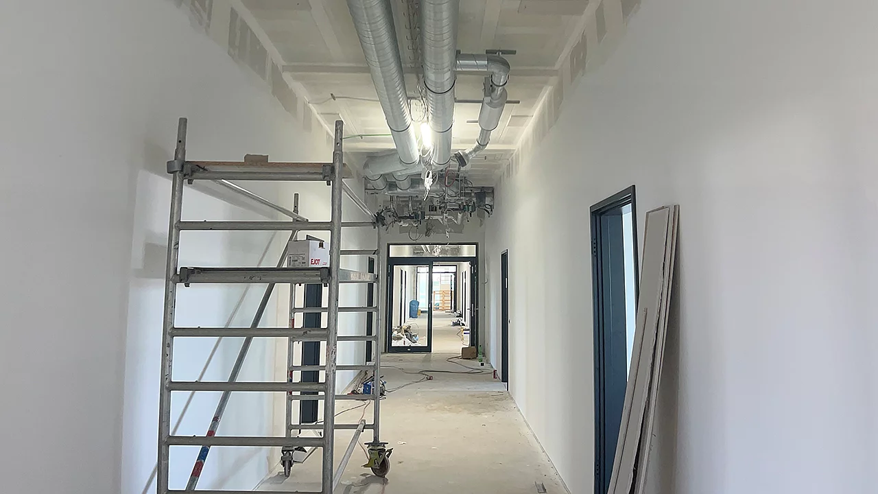 Flur eines Schulgebäudes im Innenausbau mit frisch gestrichenen Wänden, sichtbarer Lüftungstechnik unter der Decke und Arbeitsgerüst.