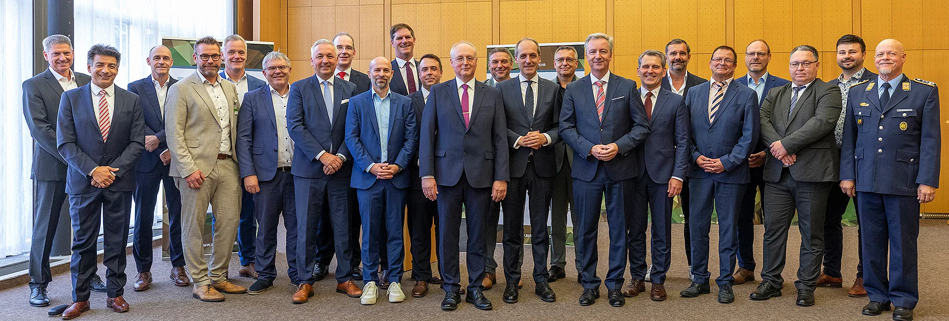 Gruppenfoto mit zahlreichen Teilnehmenden einer Veranstaltung der Bundeswehr in einem Konferenzraum. Die Personen stehen in mehreren Reihen vor Roll-ups mit Bundeswehr-Branding. Die meisten Teilnehmenden tragen Business-Kleidung, eine Person trägt eine Uniform. Im Hintergrund sind bodentiefe Fenster mit Vorhängen sowie holzverkleidete Wände zu sehen. Das Foto dokumentiert ein gemeinsames Treffen im Rahmen eines Projekts oder Vergabeverfahrens der Bundeswehr.