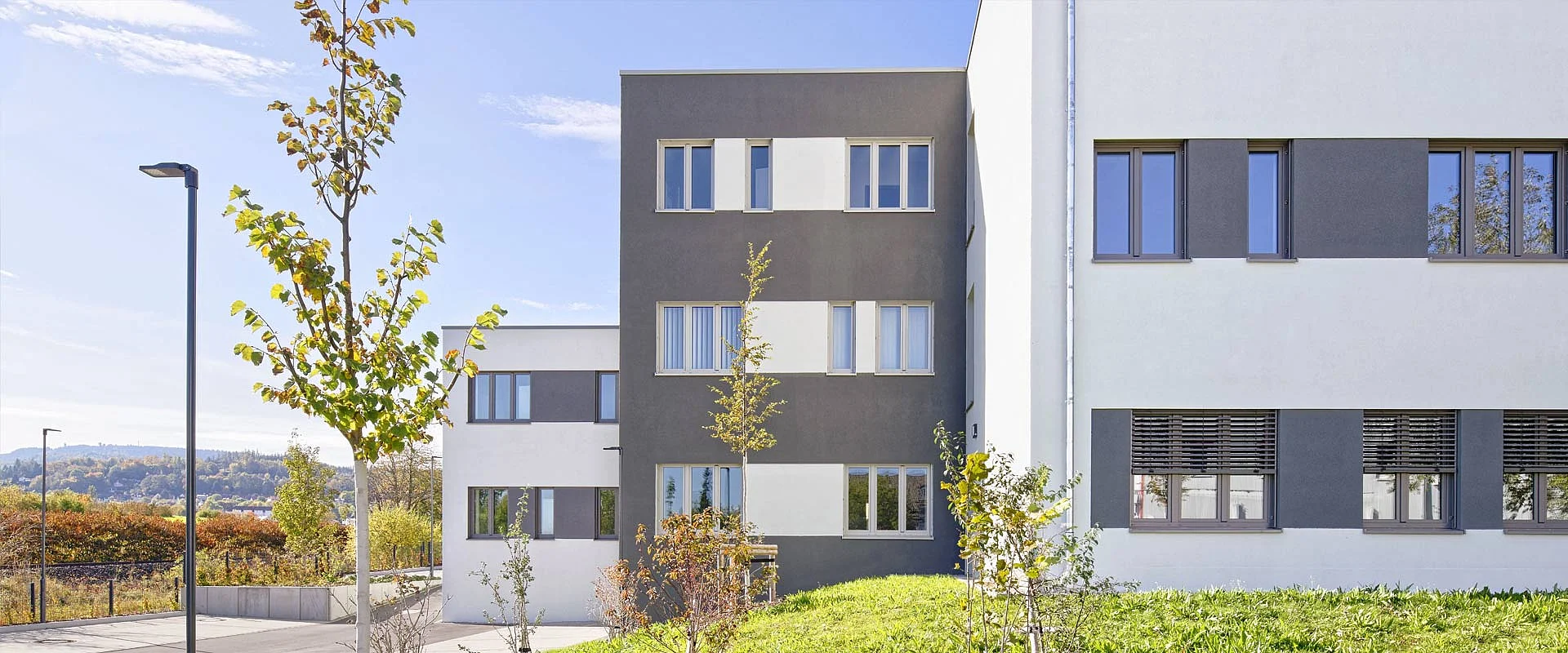 Ein modernes Gebäude mit grauen und weißen Fassaden, daneben stehen junge Bäume, im Hintergrund eine hügelige Landschaft unter klarem, blauem Himmel.