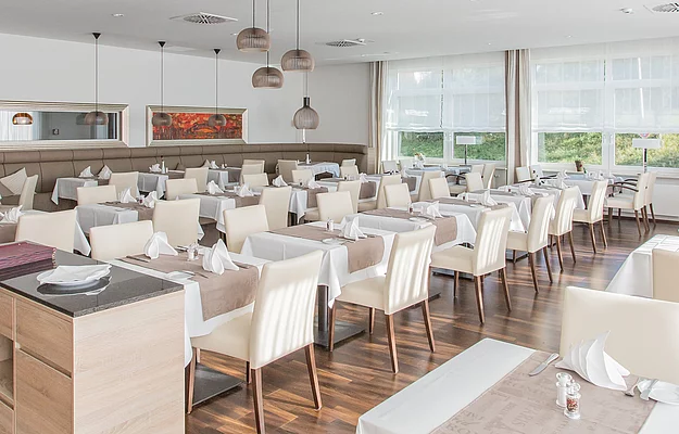 Ein eleganter Speisesaal mit weiß gedeckten Tischen, hellen Stühlen und modernen Lampen. Große Fenster bieten Ausblick auf eine grüne Landschaft.