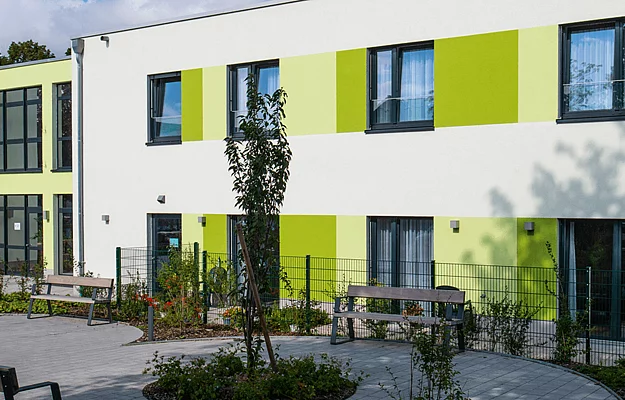 Ein modernes Gebäude mit hellgrünen Akzenten steht in einem gepflasterten Hof mit Bänken und Pflanzenbeeten, von einem hohen Zaun umgeben.