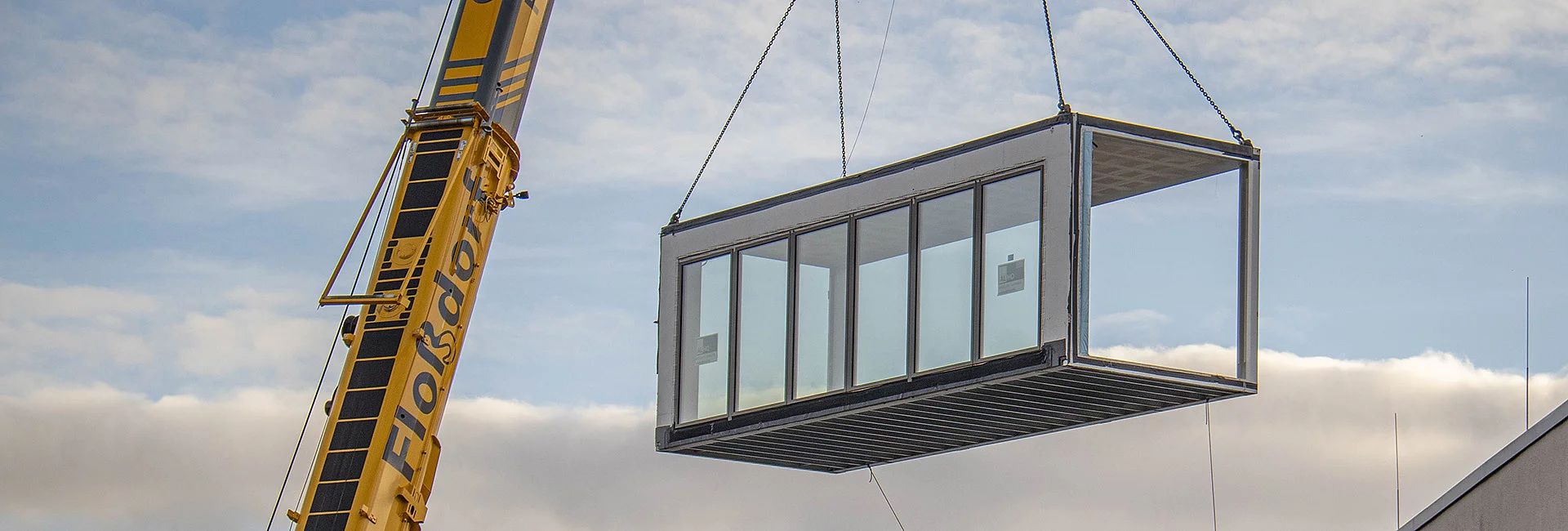 Ein gelber Kran hebt ein großes, rechteckiges Modul mit Glaswänden in die Luft. Der Himmel ist bewölkt. Auf dem Kran steht: "Wolffdorf".