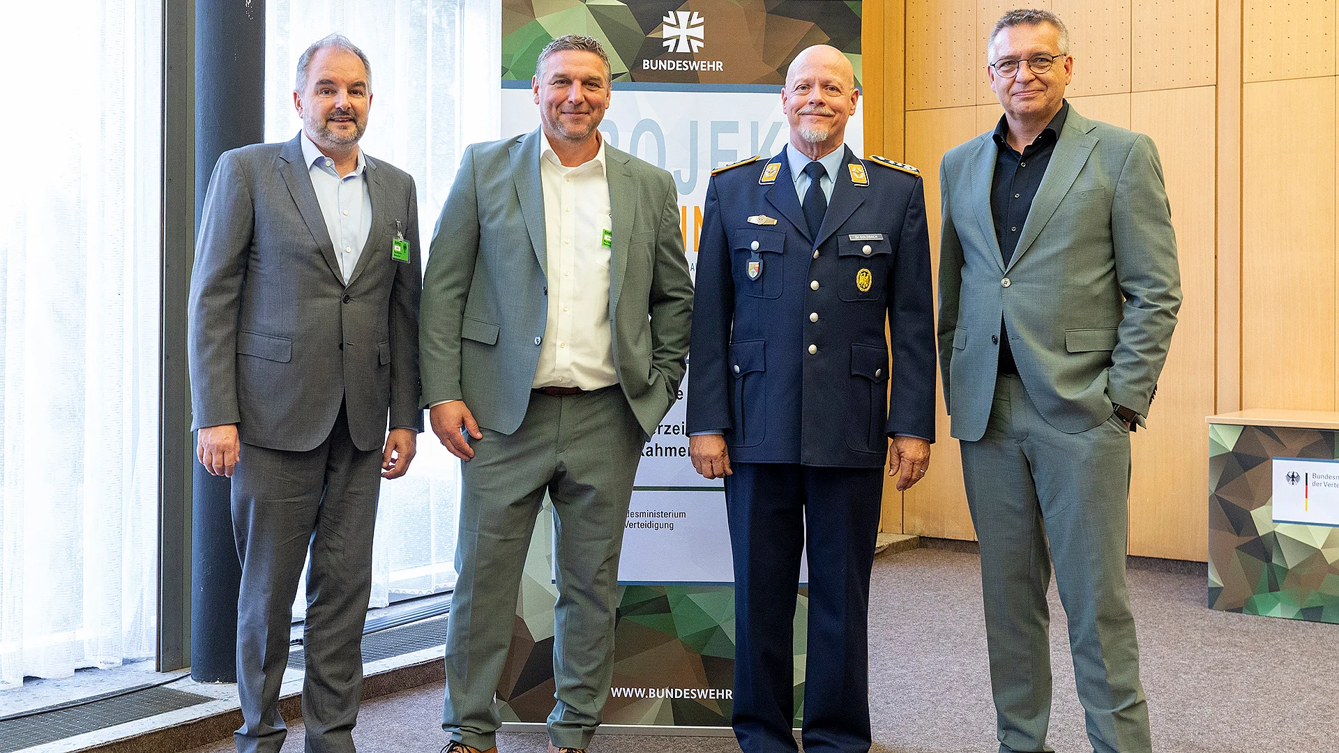 Vier Personen stehen in einem Konferenzraum vor einem Roll-up der Bundeswehr. Eine Person trägt eine Uniform, die drei anderen sind in Business-Kleidung gekleidet. Im Hintergrund sind bodentiefe Fenster mit Vorhängen sowie eine Holzverkleidung an der Wand zu sehen. Das Gruppenfoto entstand im Rahmen einer Veranstaltung der Bundeswehr.