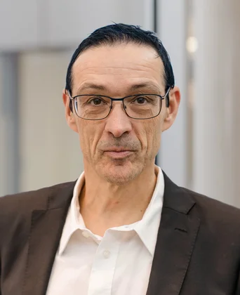 Porträt eines Mannes mit Brille im Innenraum, dunkel gekleidet mit Sakko und weißem Hemd, neutraler Gesichtsausdruck, unscharfer moderner Hintergrund.