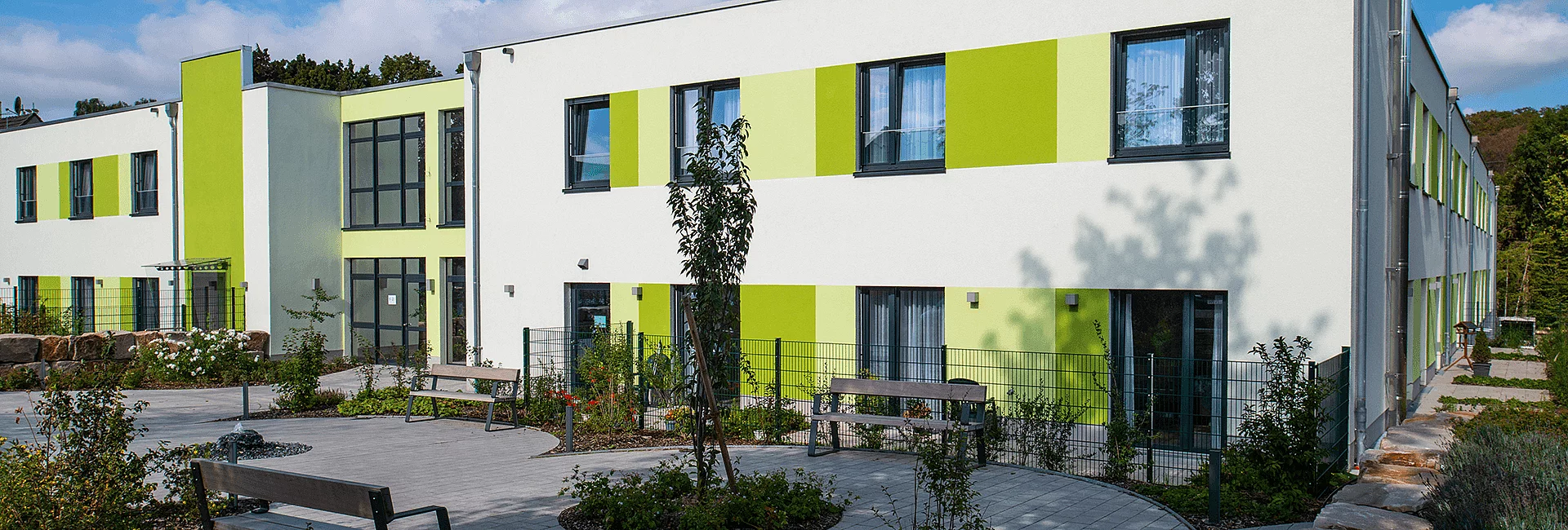 Ein modernes, zweistöckiges Gebäude mit flachem Dach und grün-weißer Fassade umrahmt einen Innenhof. Ringsum gepflegte Grünflächen und ein Parkplatz mit mehreren Autos.