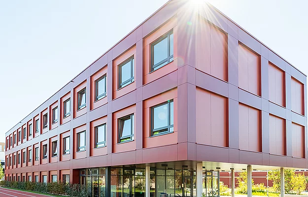 Ein rotes, modernes dreistöckiges Gebäude mit großen Fenstern steht bei Sonnenschein in einer urbanen Umgebung, umgeben von Bäumen und einem Parkplatz.