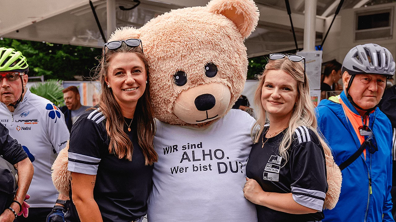 ALHO-Maskottchen posiert zusammen mit zwei Teilnehmerinnen bei einer Veranstaltung im Freien.