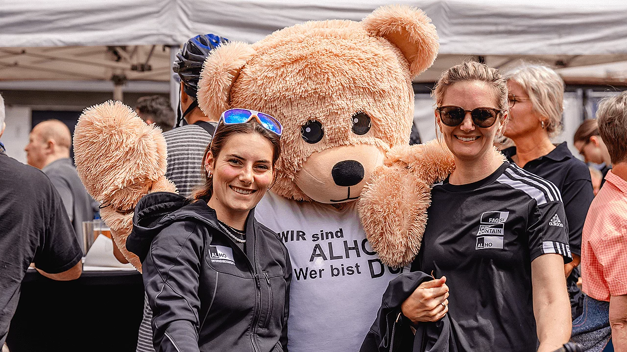 Zwei Teilnehmerinnen posieren gemeinsam mit dem ALHO-Maskottchen auf dem Veranstaltungsgelände.