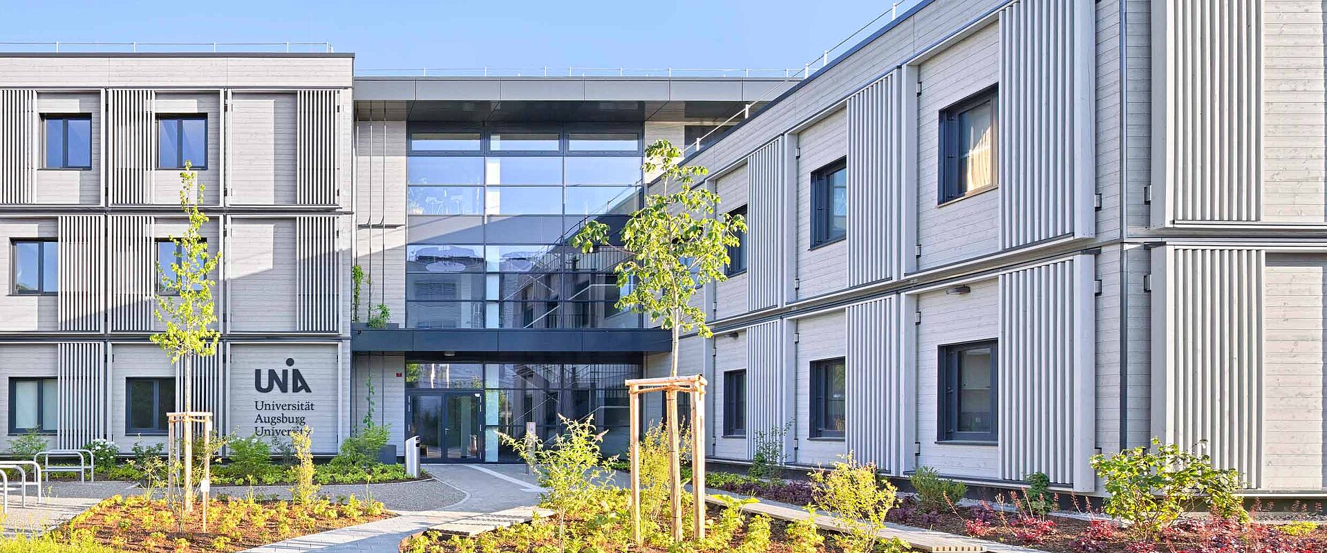 Gebäude mit modernen vertikalen Lamellen, Glasfassade im Zentrum, umgeben von grünen Pflanzen und kleinen Bäumen. Beschriftung: "UNI, Universität Augsburg University" auf der linken Seite.