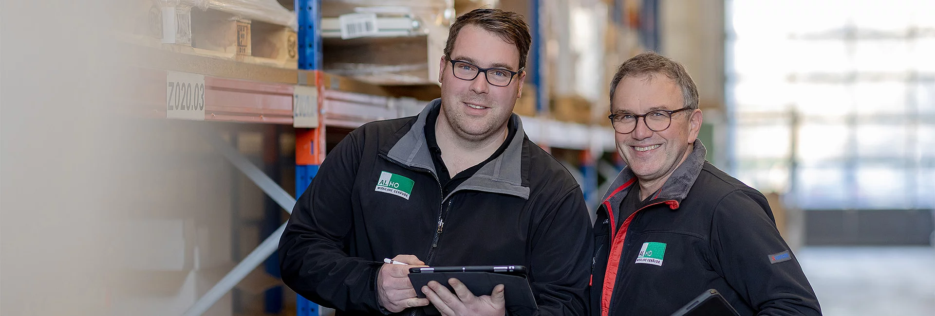 Zwei Mitarbeiter in Lagerhalle, beide mit ALHO-Jacken und Tablets, lächelnd vor Regalen mit gelagerter Ware.