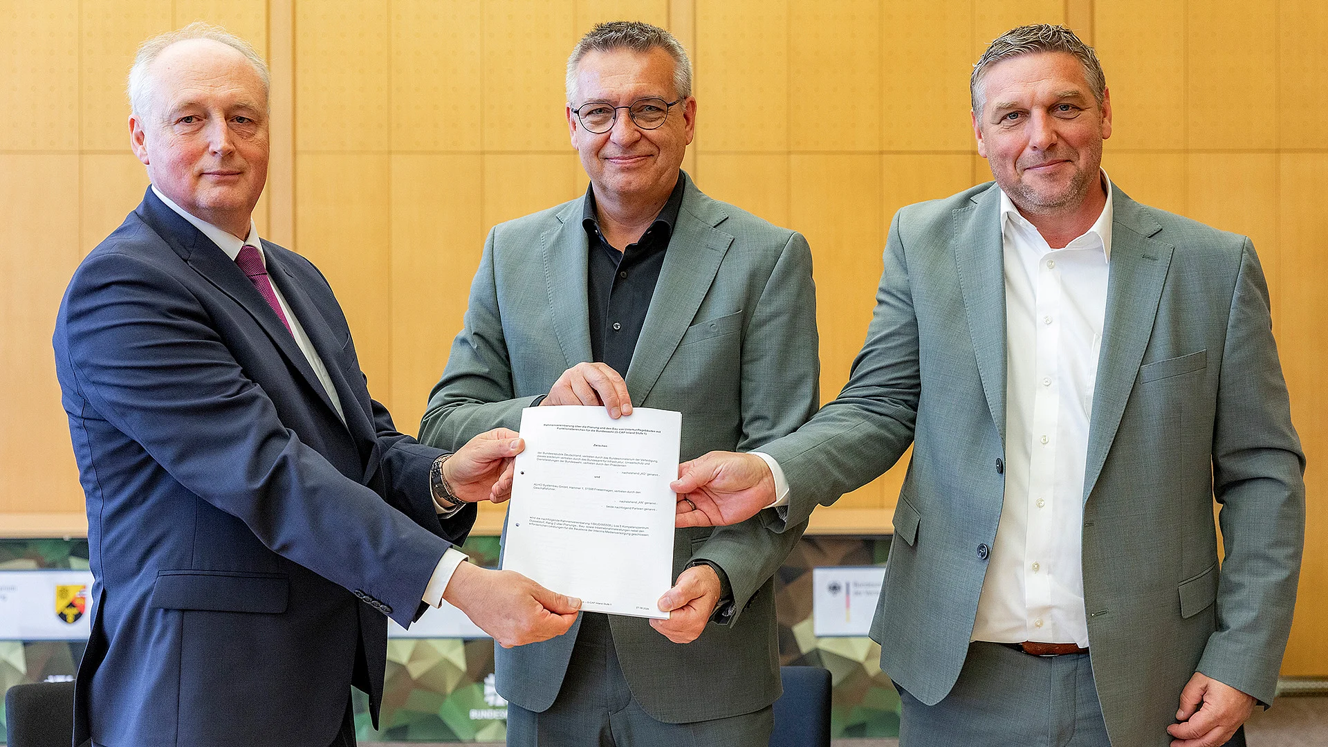 Drei Personen in Businesskleidung stehen in einem Besprechungsraum und halten gemeinsam ein Dokument, das an die Kamera präsentiert wird.