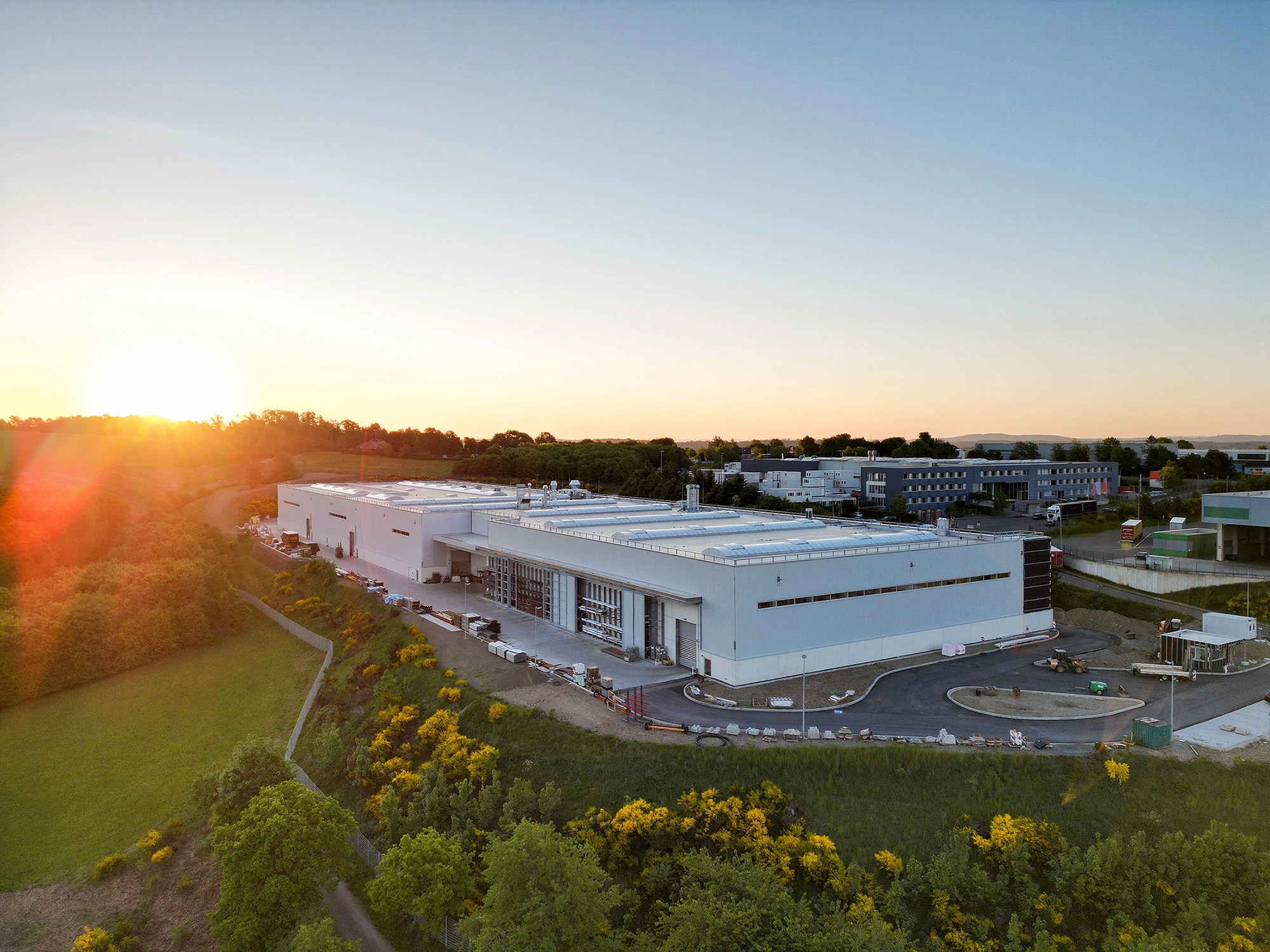 Ein großes, modernes Industriegebäude steht in einer grünen Landschaft, umgeben von Bäumen und Büschen, während die Sonne am Horizont untergeht.