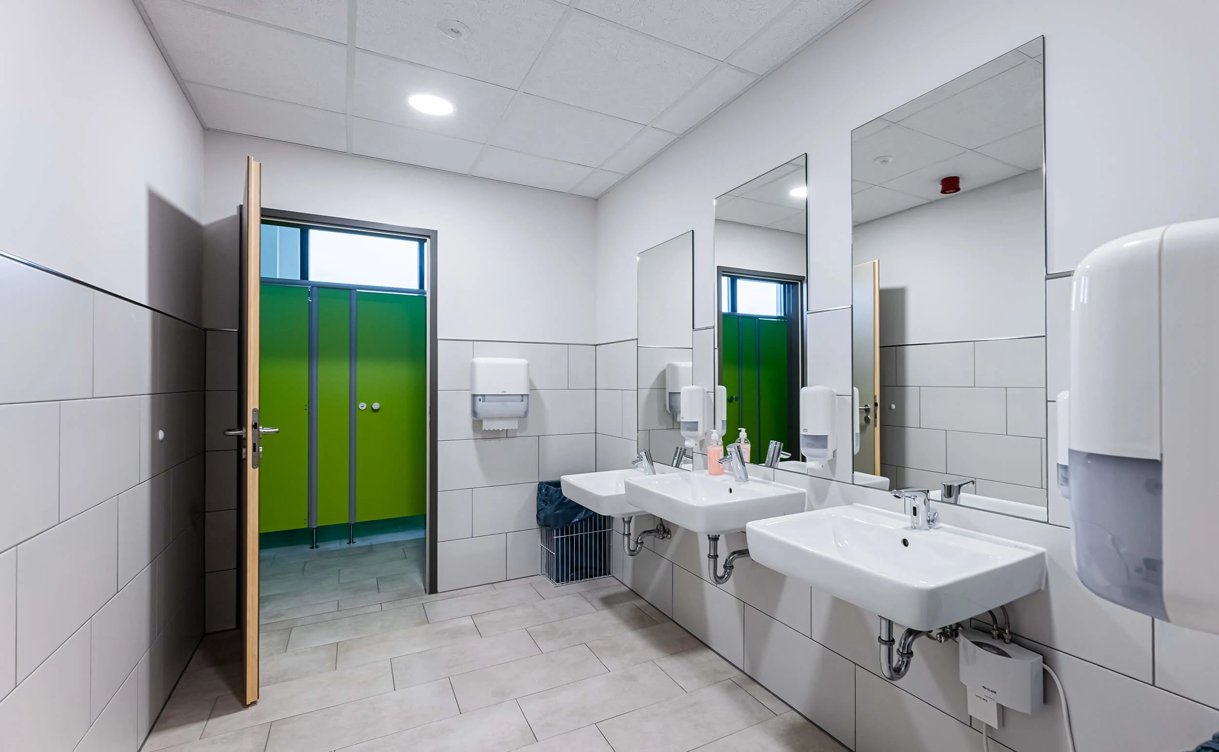Waschraum mit zwei Waschbecken, großen Spiegeln, Seifenspendern und grünen WC-Kabinen