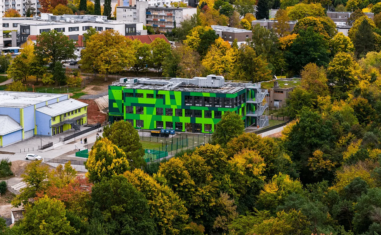 Luftaufnahme eines grünen Schulgebäudes mit Sportplatz, Bäumen und Wohnbebauung im Umfeld