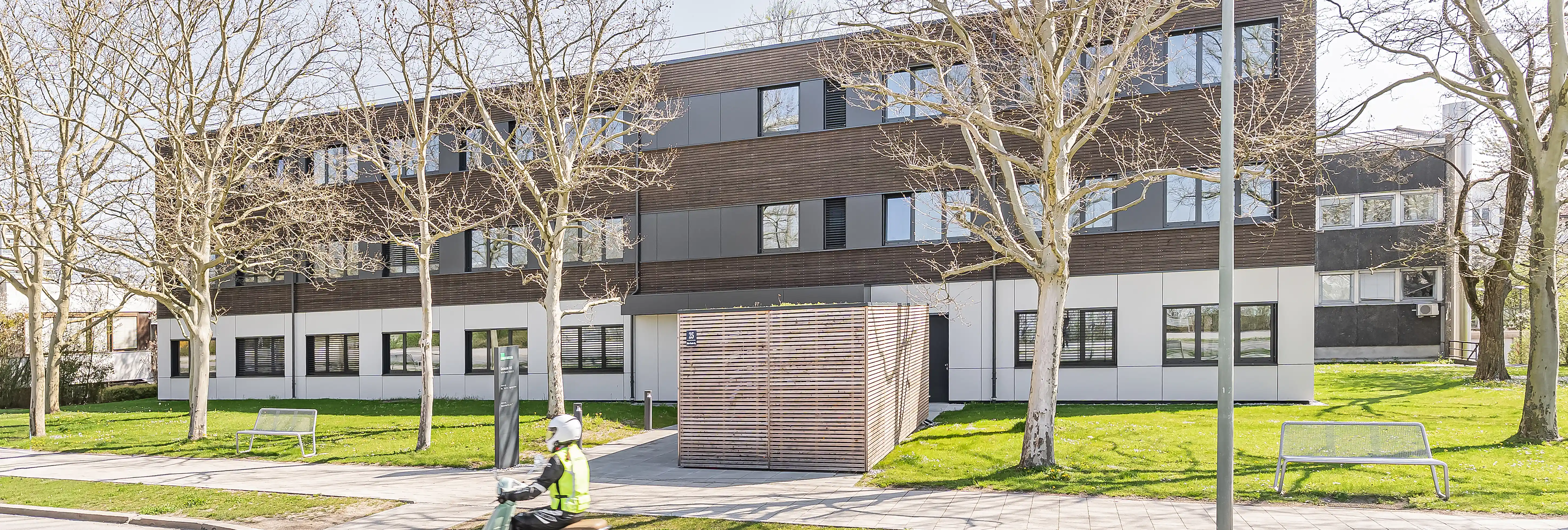 Dreigeschossiges Modulgebäude mit Holzlamellen-Fassade und hellem Sockel, eingebettet in parkähnliche Umgebung mit kahlen Bäumen und Rasenflächen