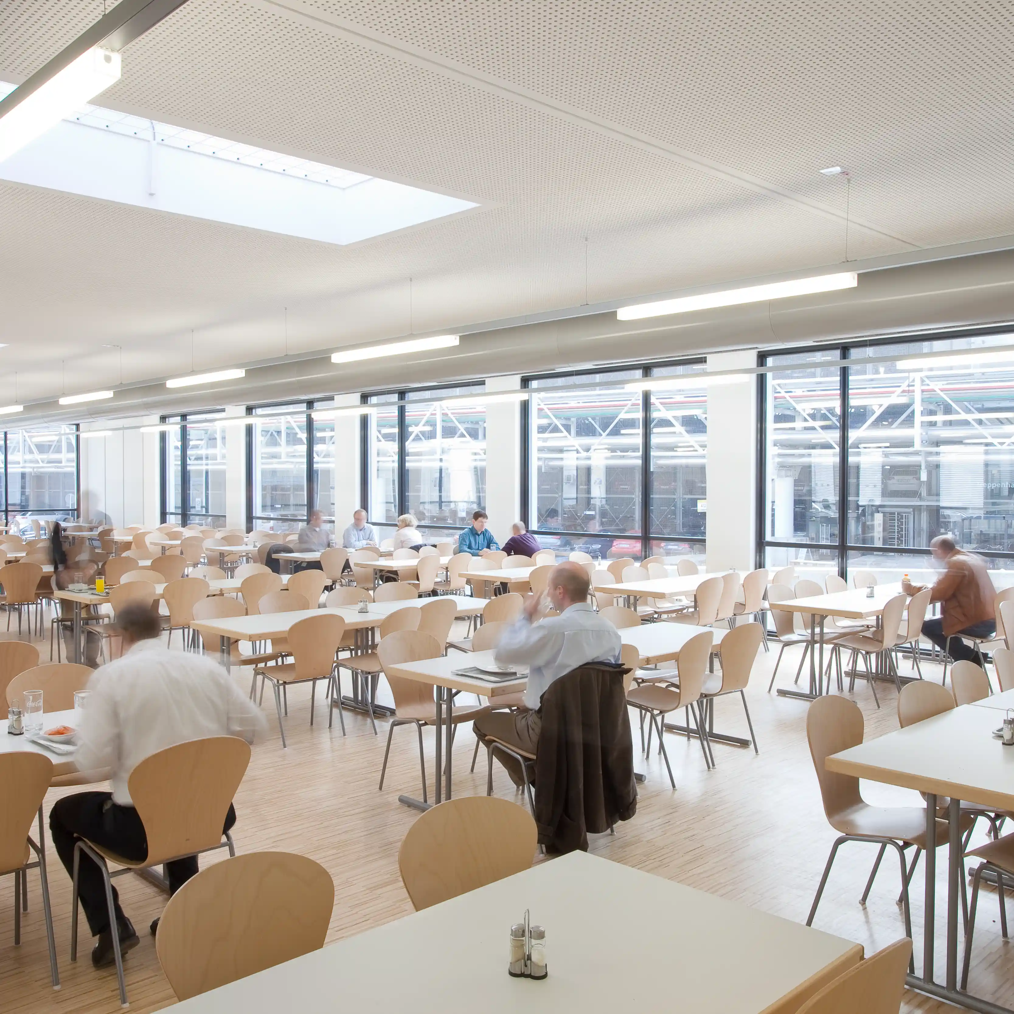 Kantine mit vielen leeren Tischen und Stühlen. Einige Personen sitzen, unterhalten sich. Die Umgebung ist hell und modern mit großen Fenstern und weißen Wänden.