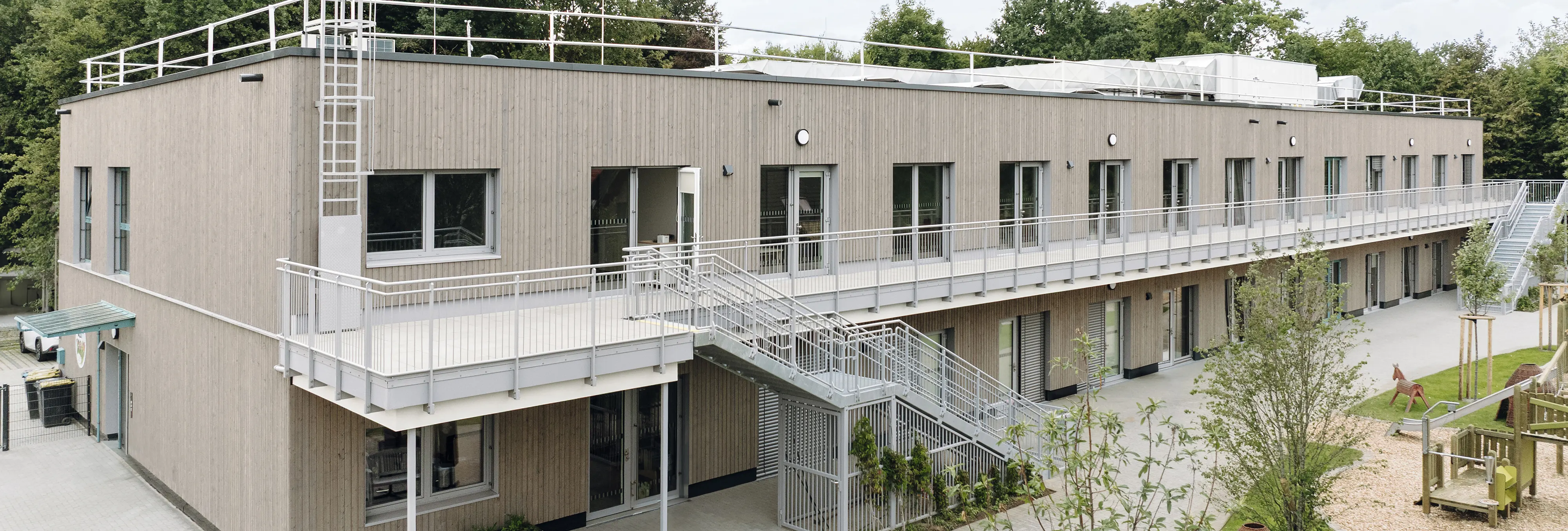 Zweigeschossiges Holzmodulgebäude mit Außentreppe, Laubengang, Spielplatz und begrüntem Außenbereich