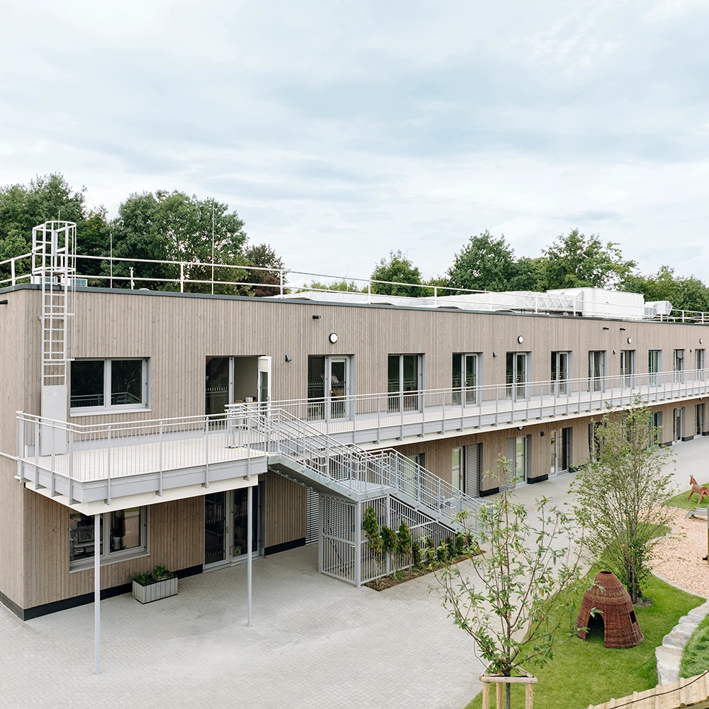 Zweigeschossiges Holzmodulgebäude mit Außentreppen, Dachtechnik und begrüntem Außenbereich