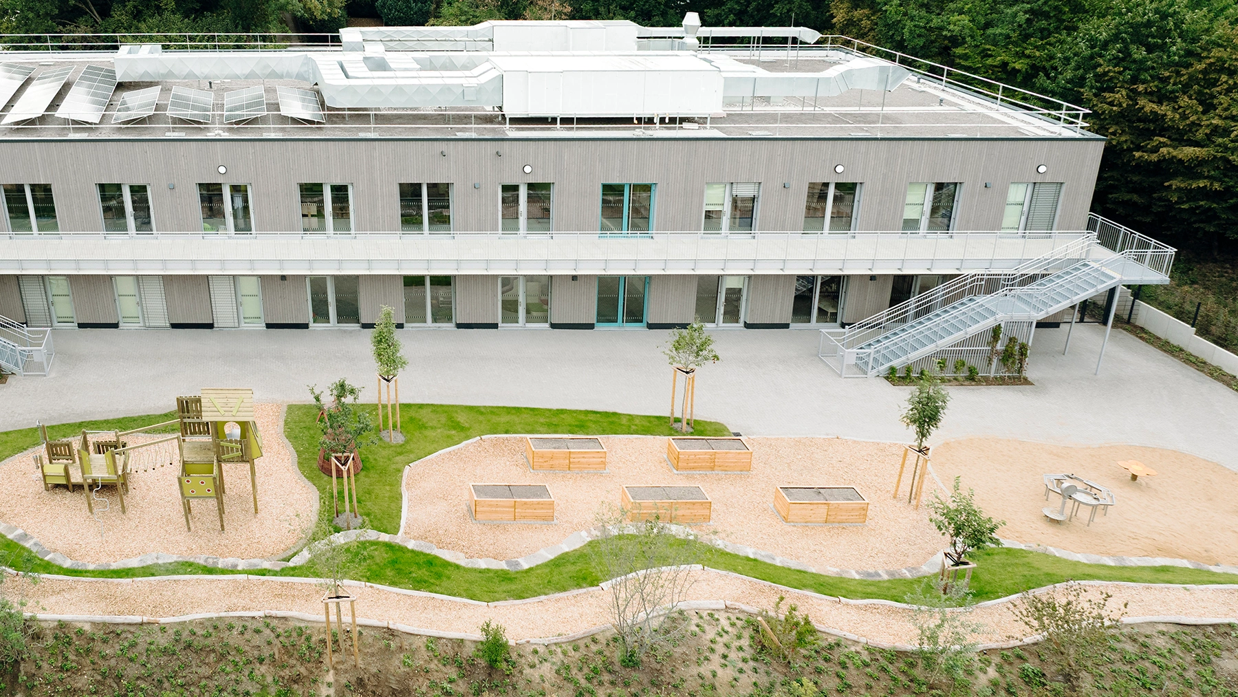 Zweigeschossiges Holzmodulgebäude mit Spielplatz, Sandflächen, Hochbeeten und Außentreppen