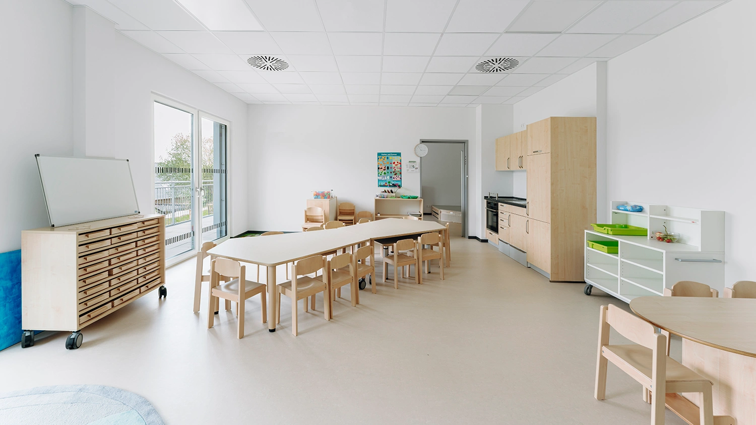 Heller Gruppenraum mit Kindertischen, kleinen Stühlen, Küchenzeile und Spielbereich