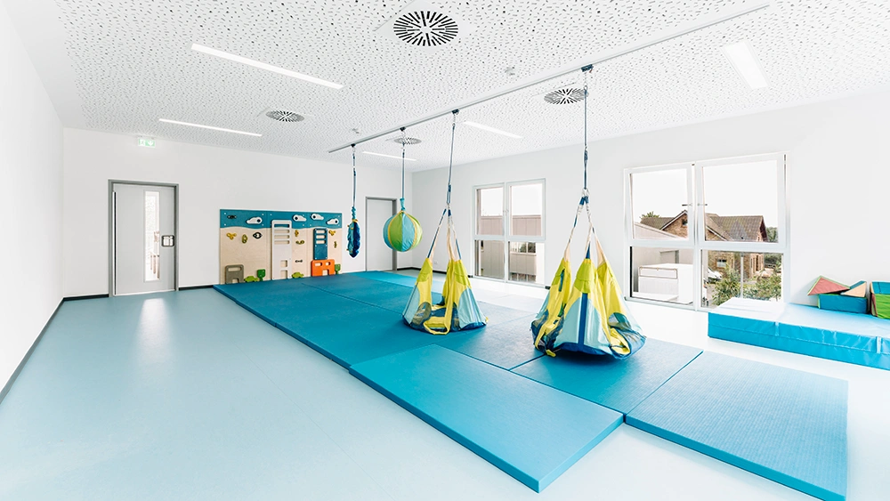 Bewegungsraum mit blauen Matten, Hängesitzen, Therapieschaukel und großen Fenstern