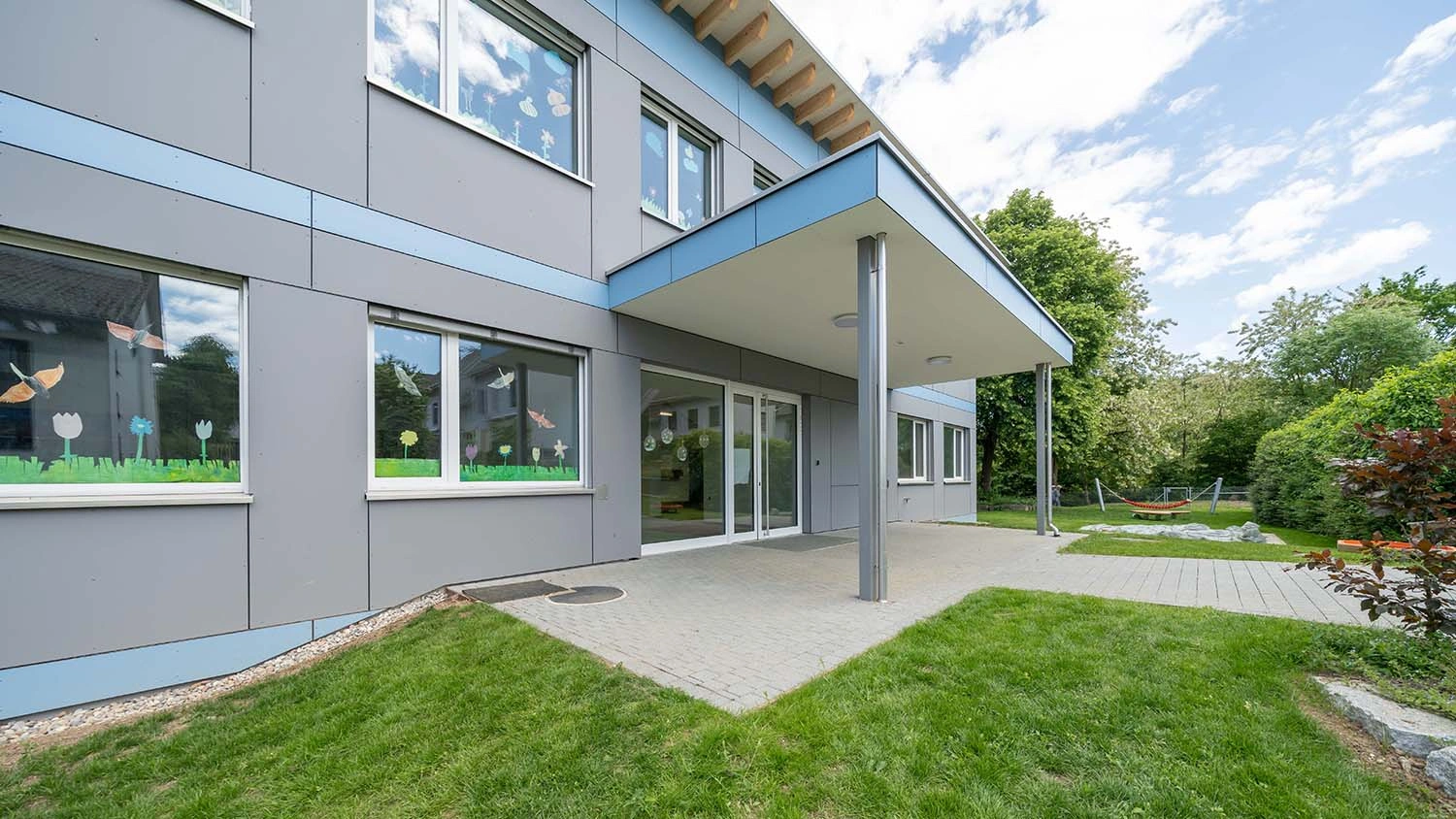 Gebäude mit grauer Fassade und großer Fensterfront, dekoriert mit Blumenbildern. Der Eingangsbereich ist überdacht, umgeben von grünem Rasen und Bäumen, mit einer Hängematte im Hintergrund.