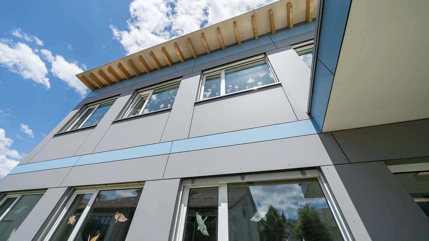 Gebäude mit moderner, grauer Fassade; bunte Papierdekorationen an den Fenstern. Der Himmel ist teils bewölkt, strahlt Blau. Holzsparren ragen aus dem Dach hervor.
