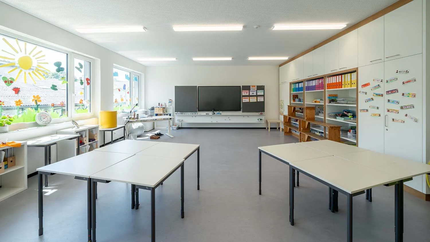 Klassenzimmer mit gruppierten Tischen; an den Fenstern bunte Dekorationen, links Regale mit Materialien; im Hintergrund ein Smartboard und Schränke mit farbigen Ordnern.