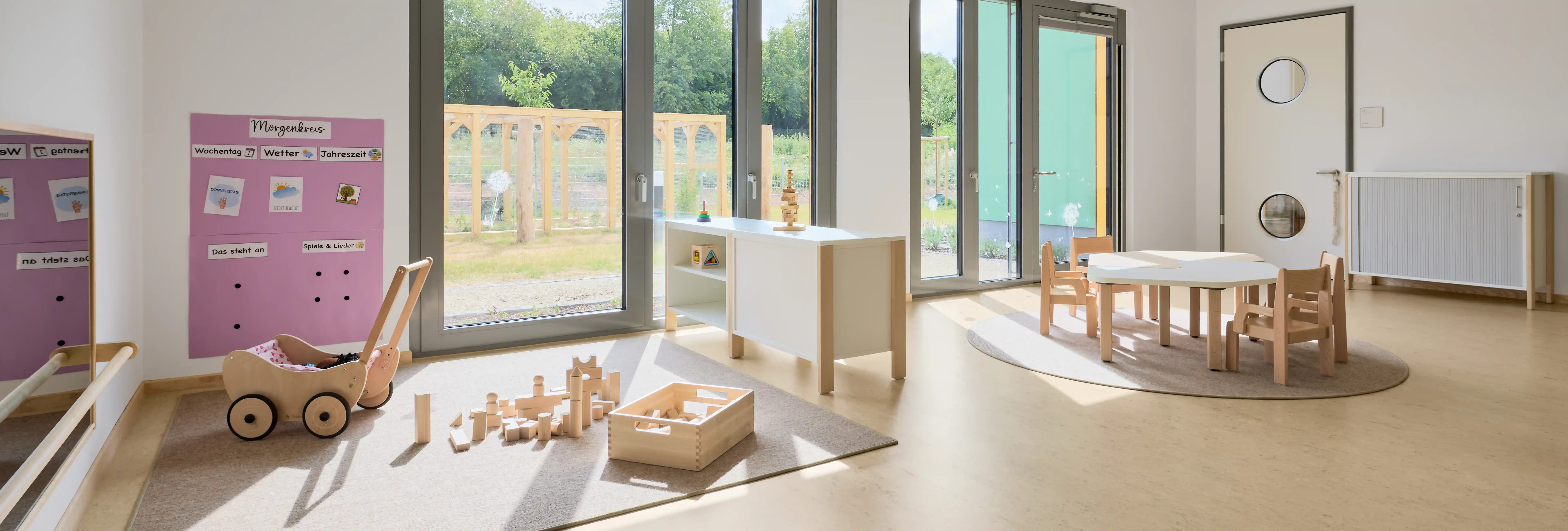 Heller, modern eingerichteter Gruppenraum einer Kindertagesstätte mit bodentiefen Fenstern und Blick ins Grüne. Auf einem Teppich liegen Holzbausteine und ein Holzpuppenwagen. Rechts befindet sich ein niedriger Kindertisch mit mehreren kleinen Holzstühlen. Tageslicht durchflutet den Raum und schafft eine freundliche Lern- und Spielumgebung.