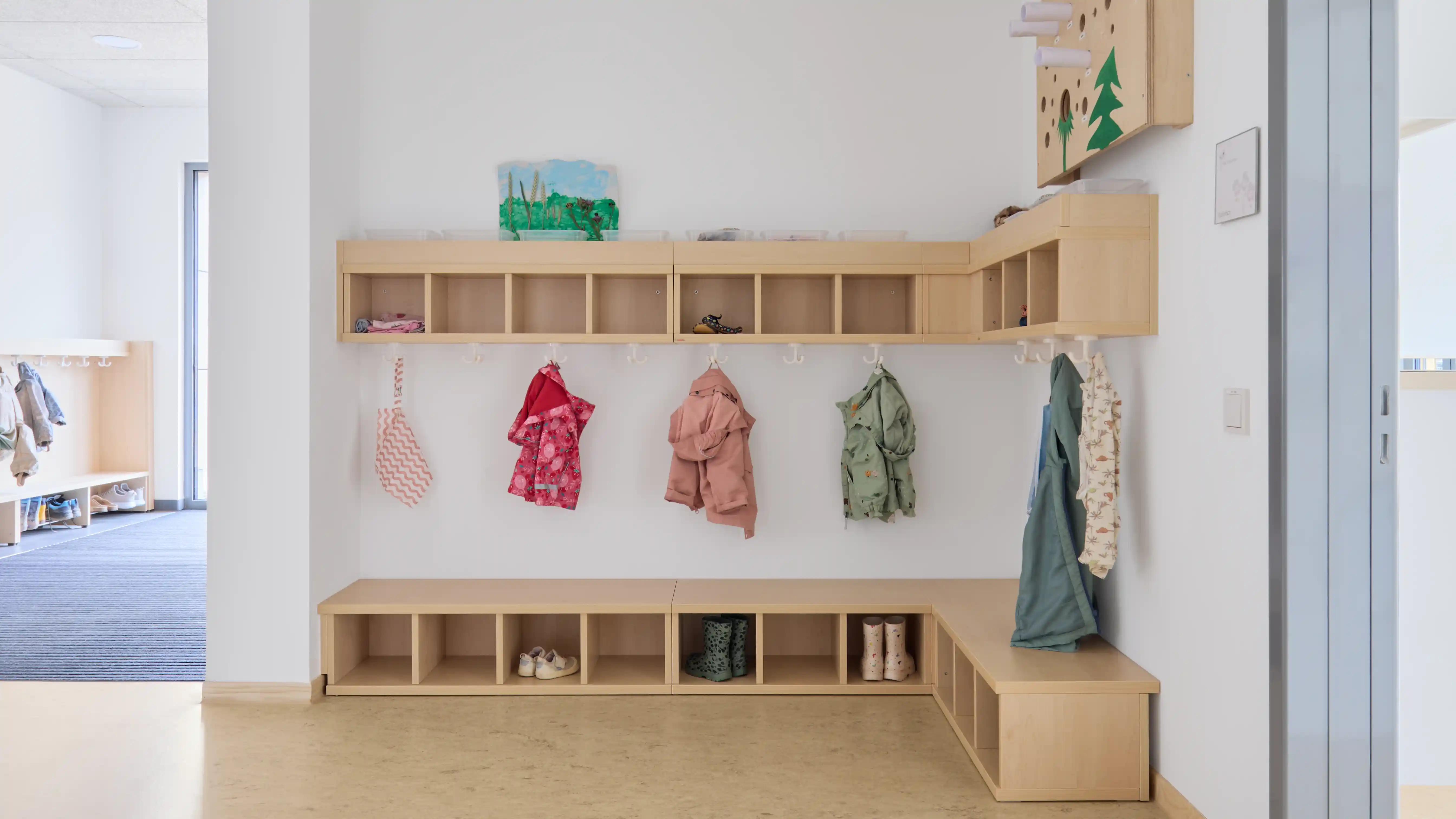 Übersichtlicher Garderobenbereich einer Kindertagesstätte mit niedrigen Holzregalen, Sitzbank und einzelnen Fächern für Kleidung und Schuhe. Mehrere Kinderjacken hängen an Haken, darüber befinden sich offene Ablagefächer. Der helle Raum ist funktional und kindgerecht gestaltet.