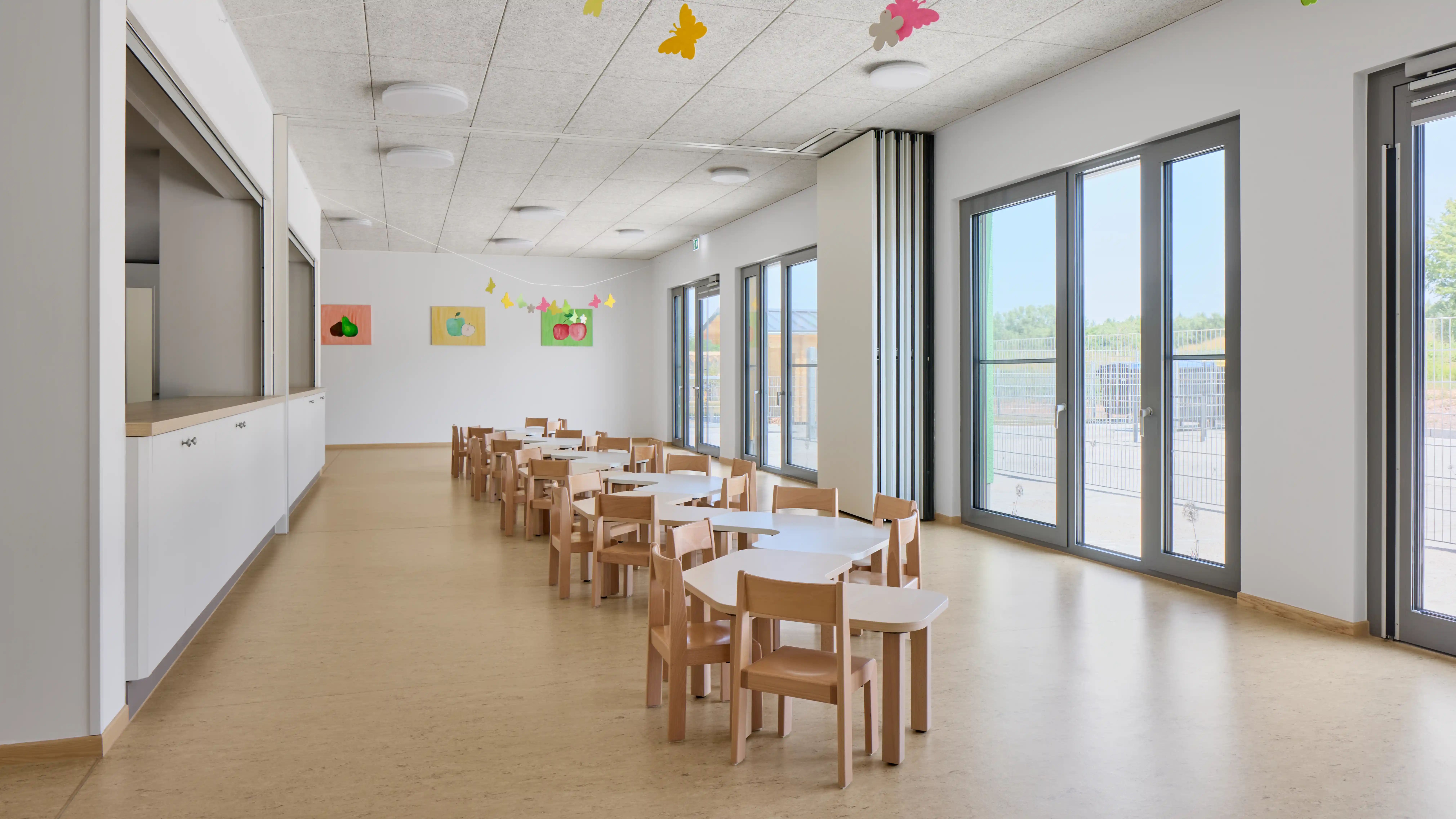 Langer, heller Mehrzweckraum mit mehreren kindgerechten Tischen und Stühlen. Bodentiefe Fenster auf der rechten Seite bieten viel Tageslicht und Ausblick nach draußen. Farbige Dekorationen und Kinderzeichnungen an den Wänden verleihen dem Raum eine freundliche und einladende Atmosphäre.