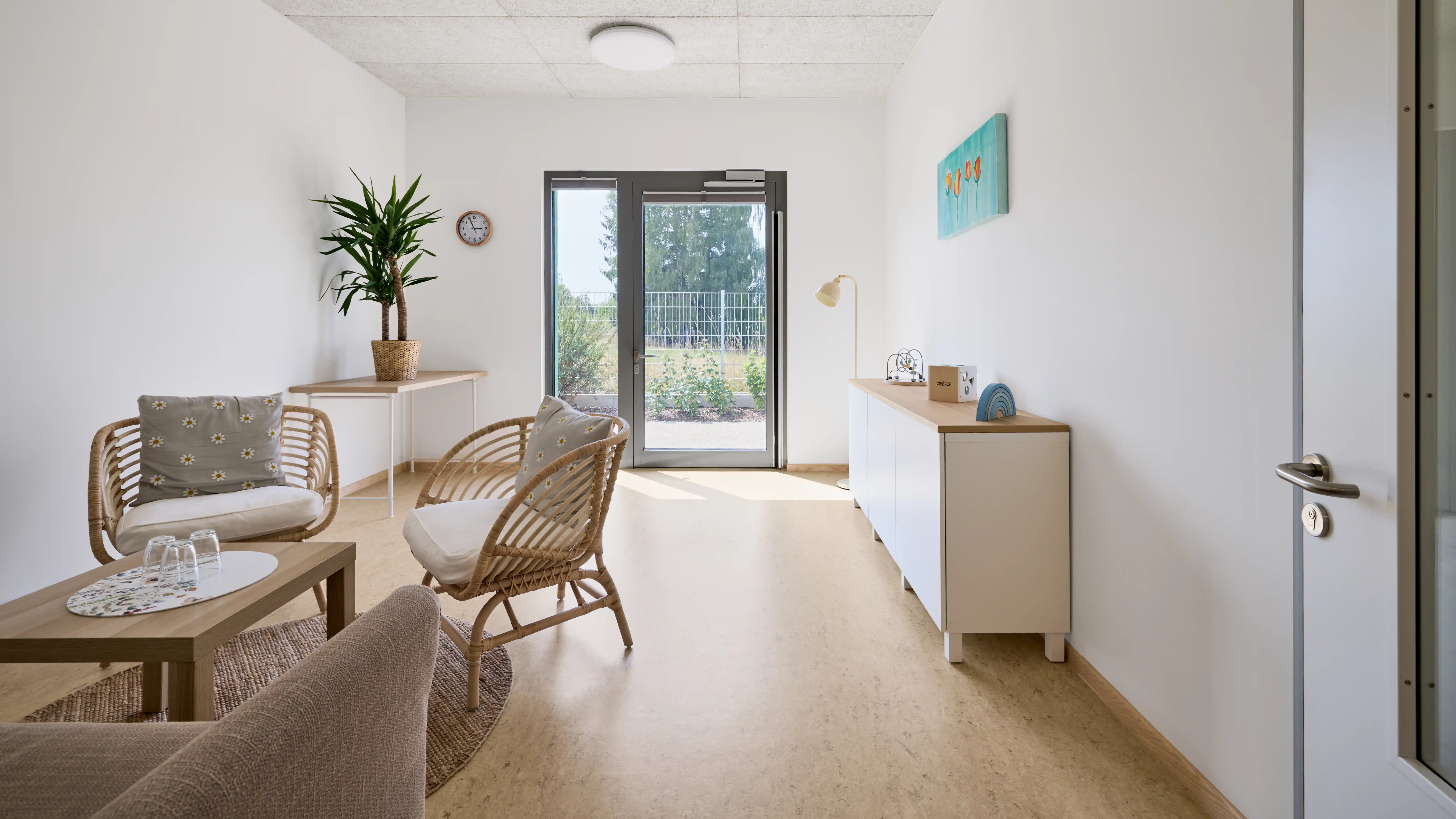 Heller, freundlich gestalteter Aufenthaltsraum mit Sitzgruppe aus Korbstühlen und Sofa um einen niedrigen Tisch. Eine Zimmerpflanze, ein Sideboard mit Spielmaterialien und eine Glastür zum Außenbereich schaffen eine ruhige und einladende Atmosphäre. Tageslicht fällt durch die Tür und beleuchtet den modern eingerichteten Raum.