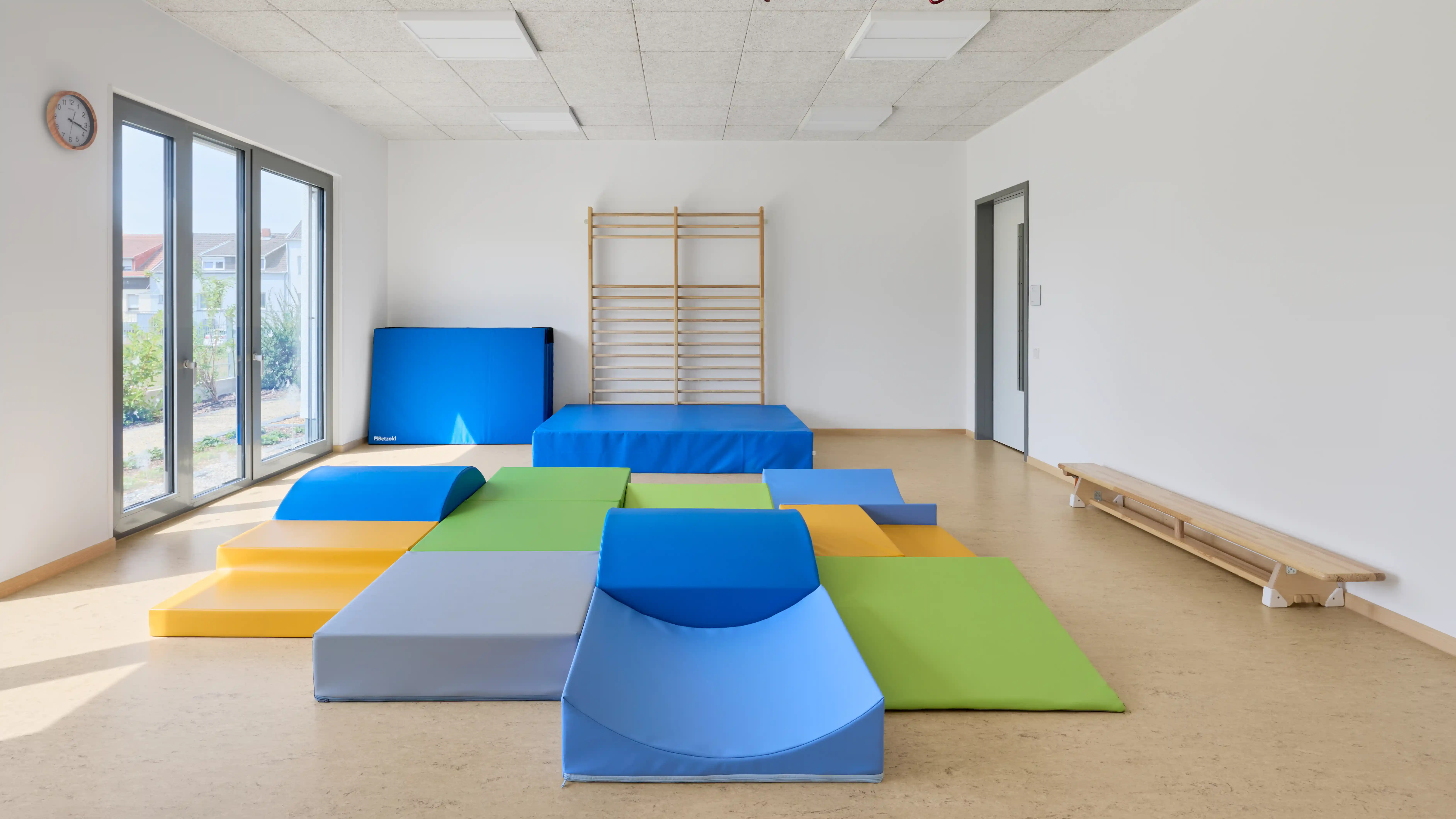 Heller Bewegungsraum in einer Kindertagesstätte mit bunten Weichboden- und Turnelementen in Blau, Grün, Gelb und Grau. An der Rückwand befindet sich eine Sprossenwand mit zusätzlichen Matten, während große Fenster auf der linken Seite viel Tageslicht in den Raum lassen. Die Ausstattung bietet vielfältige Möglichkeiten für Bewegung, Spiel und motorische Förderung.