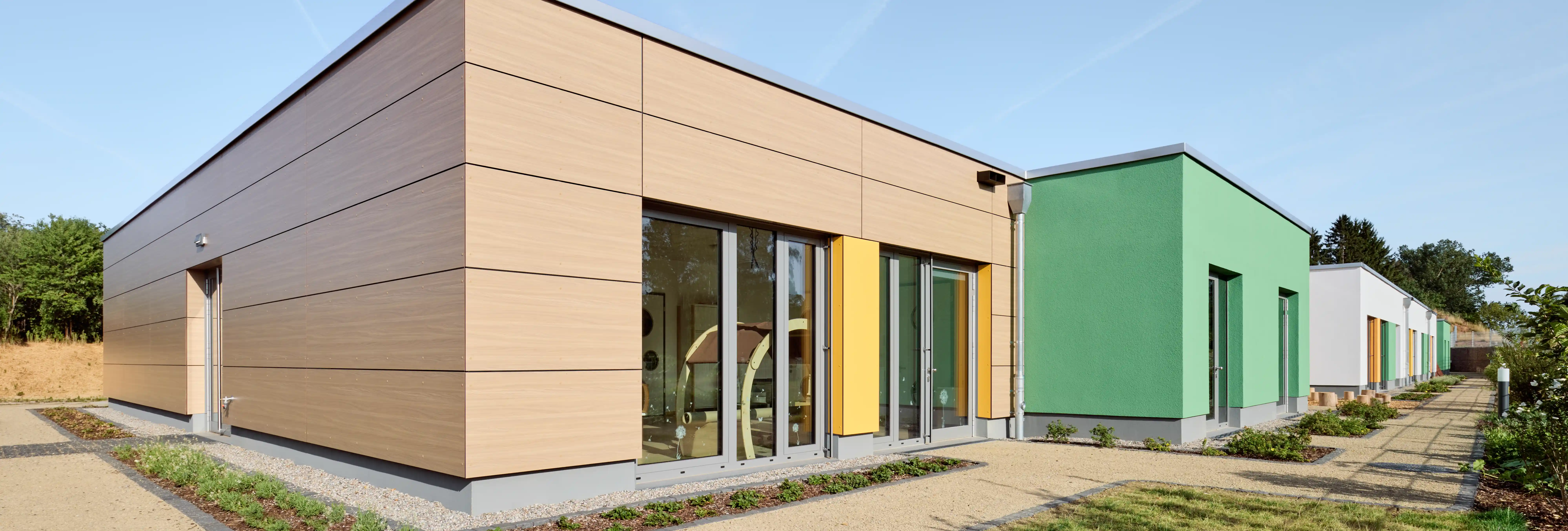 Moderne Kindertagesstätte mit einer Fassade aus Holz- und farbigen Gebäudeelementen. Große Fensterfronten sorgen für viel Tageslicht im Innenraum. Gepflasterte Wege und bepflanzte Außenflächen umgeben das Gebäude in einer gepflegten, naturnahen Umgebung.
