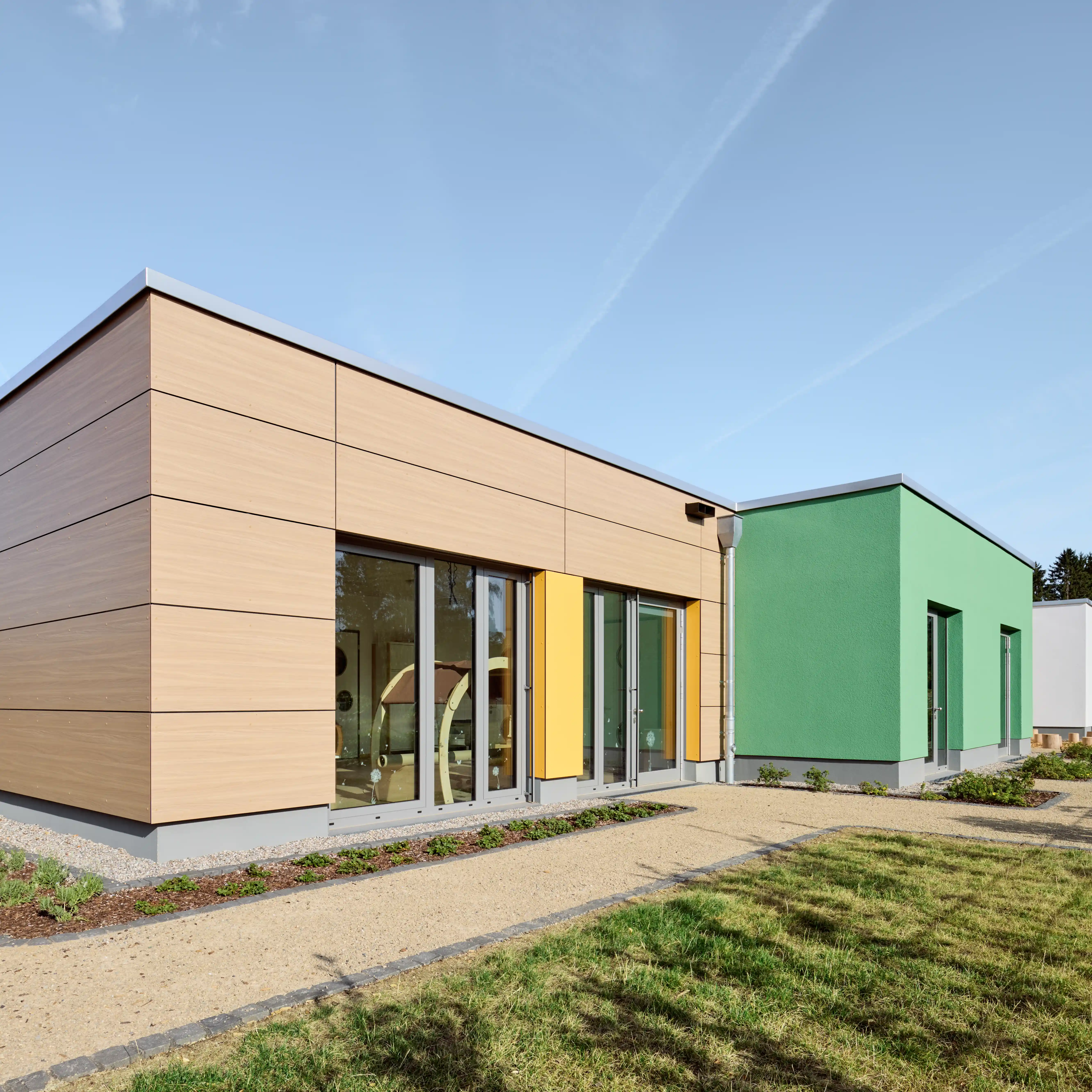 Moderne Kindertagesstätte mit einer Fassade aus Holz- und farbigen Gebäudeelementen. Große Fensterfronten sorgen für viel Tageslicht im Innenraum. Gepflasterte Wege und bepflanzte Außenflächen umgeben das Gebäude in einer gepflegten, naturnahen Umgebung.