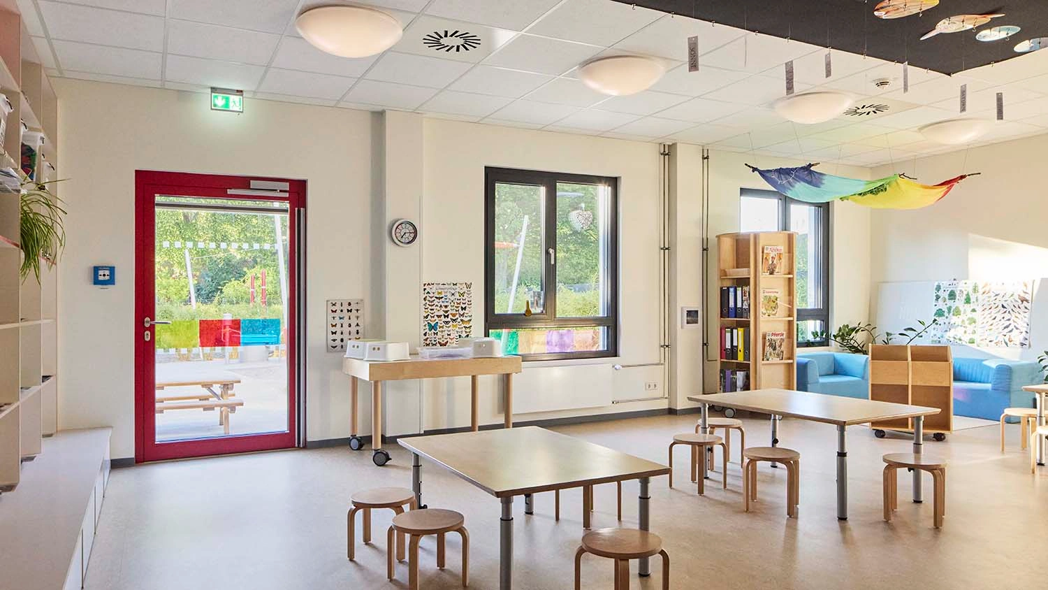 Kindertagesstätte: Mehrere Holztische und -hocker stehen in einem hellen Raum mit großen Fenstern. An der Decke hängen bunte Stoffbahnen. Regale enthalten Bücher und Pflanzen.
