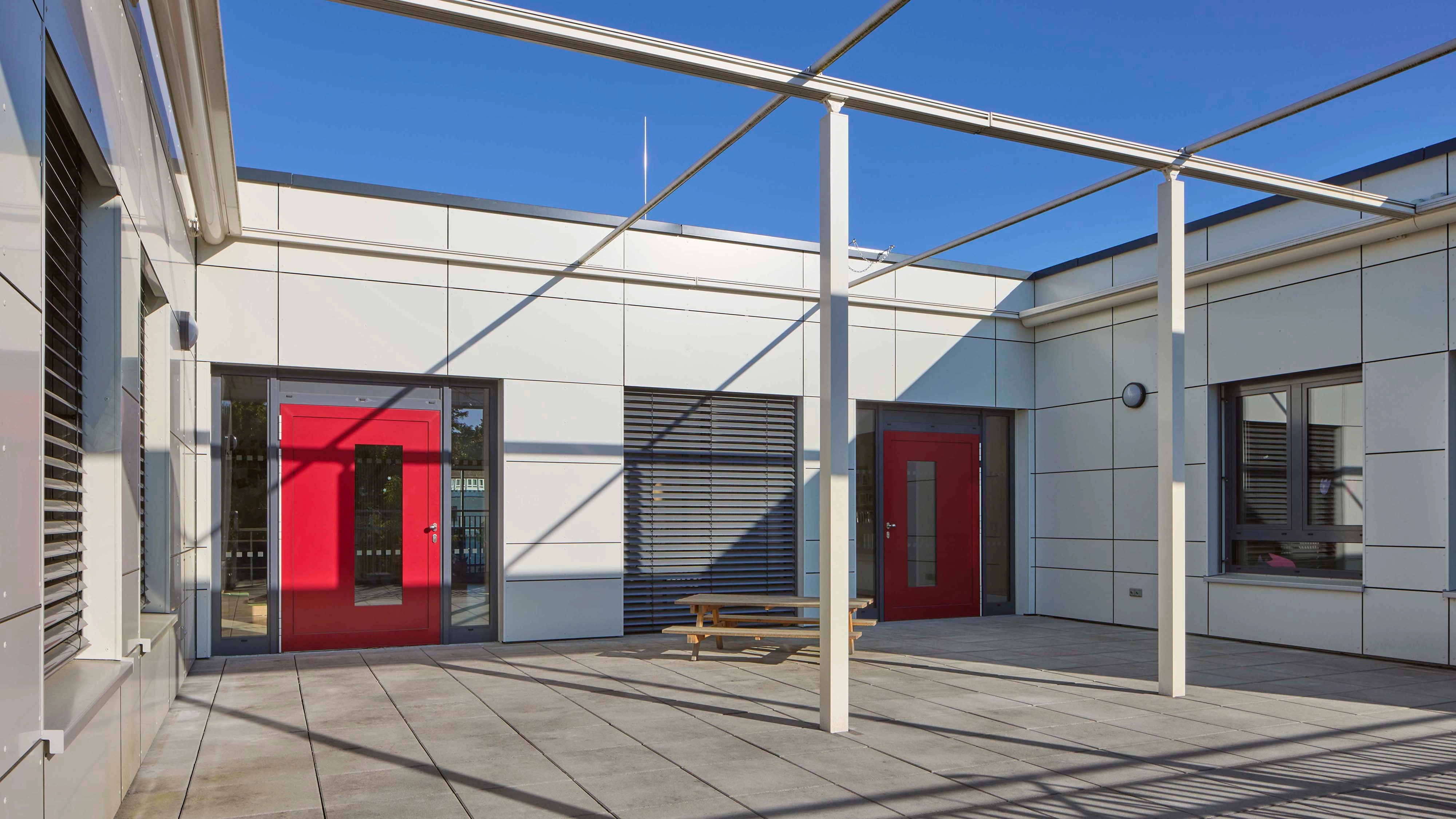 Roter Eingang in rechteckigem weißen Gebäude, Schatten geometrischer Strukturen und Holzbank im Innenhof, klare blaue Himmelsansicht.