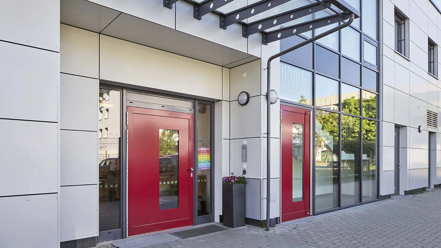 Zwei rote Glastüren öffnen sich zu einem modernen Gebäude. Daneben befindet sich ein Blumenbehälter auf einem gepflasterten Weg. Der Eingang ist von Glas- und Aluminium-Elementen umgeben.