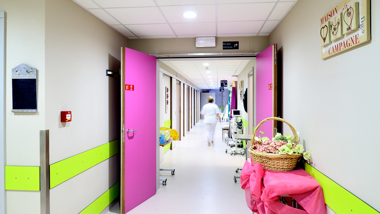 Krankenhausflur mit pink-grüner Farbgestaltung und einer Pflegekraft im Hintergrund