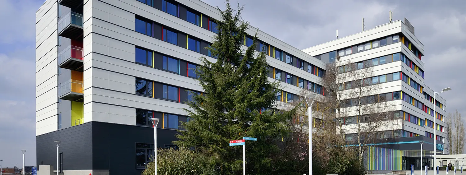 Mehrgeschossiges Klinikgebäude mit heller Fassade, farbigen Fensterelementen und Bäumen im Vordergrund
