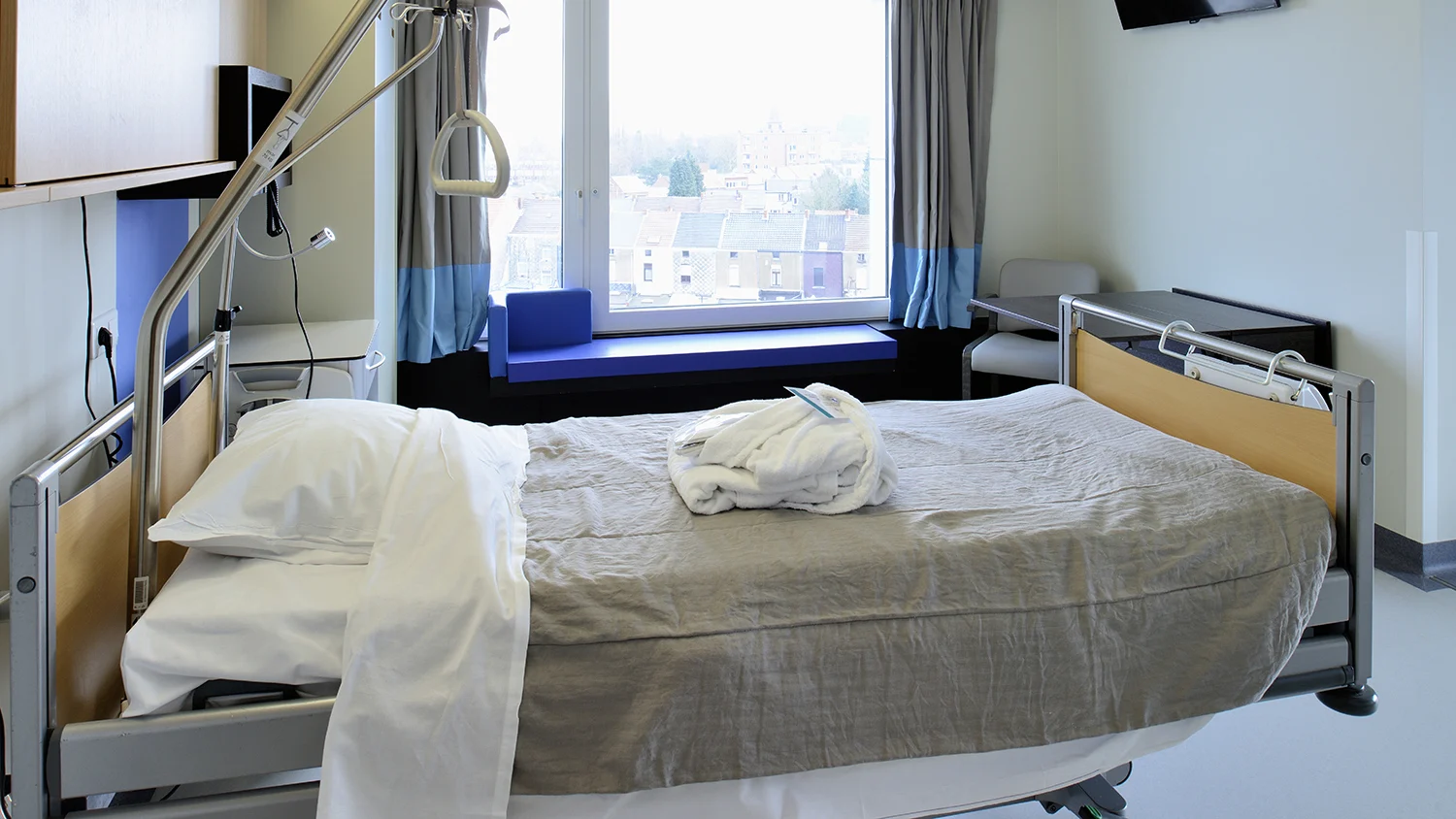 Helles Patientenzimmer mit Pflegebett, großem Fenster, Sitzbank und medizinischer Ausstattung