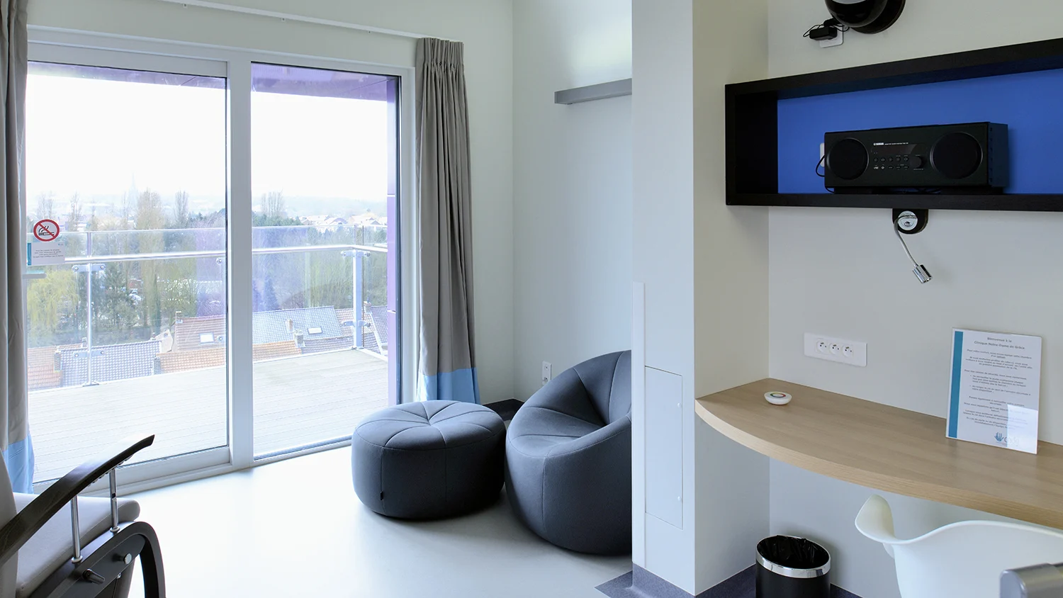 Patientenzimmer mit Sessel, Schreibtisch, Audiogerät und bodentiefem Fenster zum Balkon