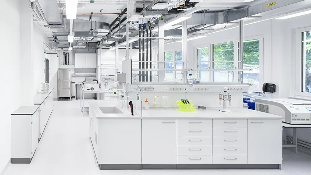 Helles Labor mit weißen Arbeitsflächen, Laborgeräten, Fenstern und sichtbarer Deckentechnik