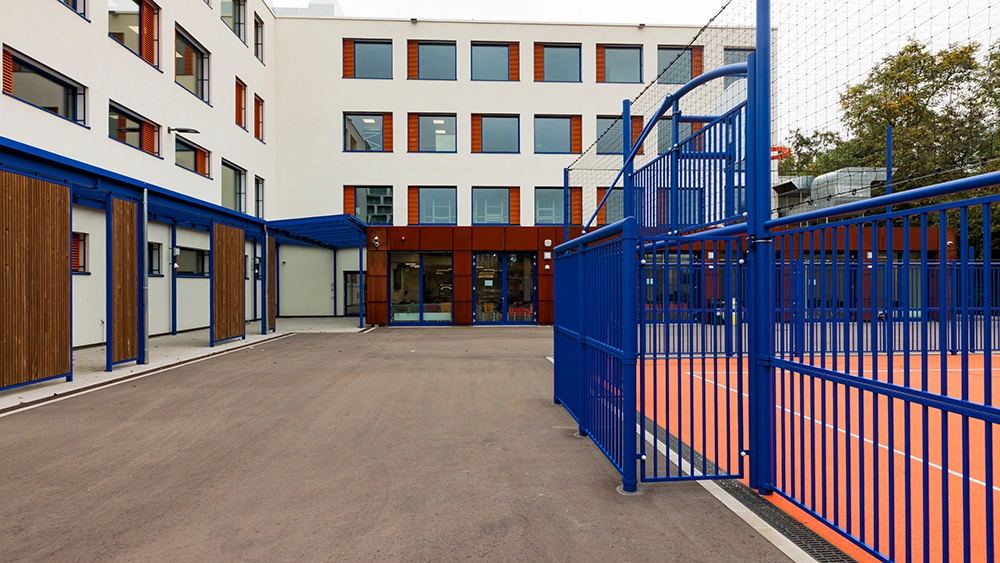 Gebäude mit weißen Wänden und roten Fensterläden, blaue Struktur mit Holzpaneelen. Rechts eingezäunter, orangefarbener Sportplatz. Im Hintergrund Bäume und Lüftungsanlage.