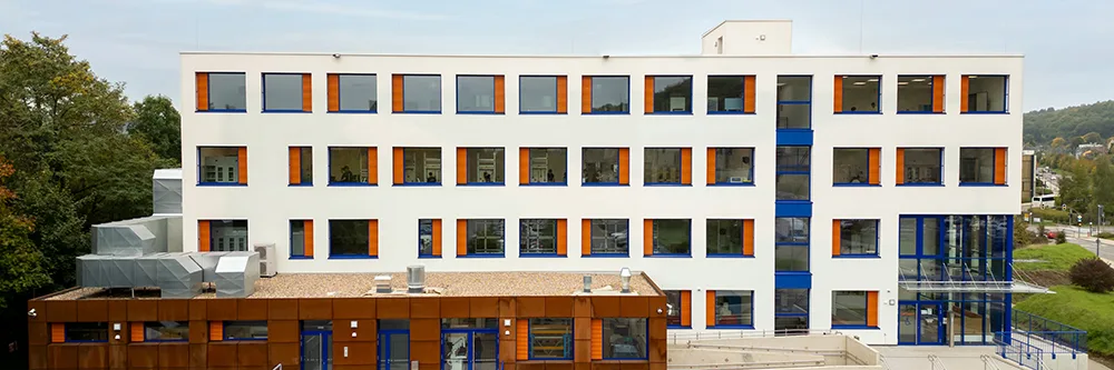 Ein modernes, vierstöckiges Gebäude mit weißen Wänden, blauen Fensterrahmen und orangefarbenen Jalousien. Es steht in einer grünen, bewaldeten Umgebung mit Straßen in der Nähe.