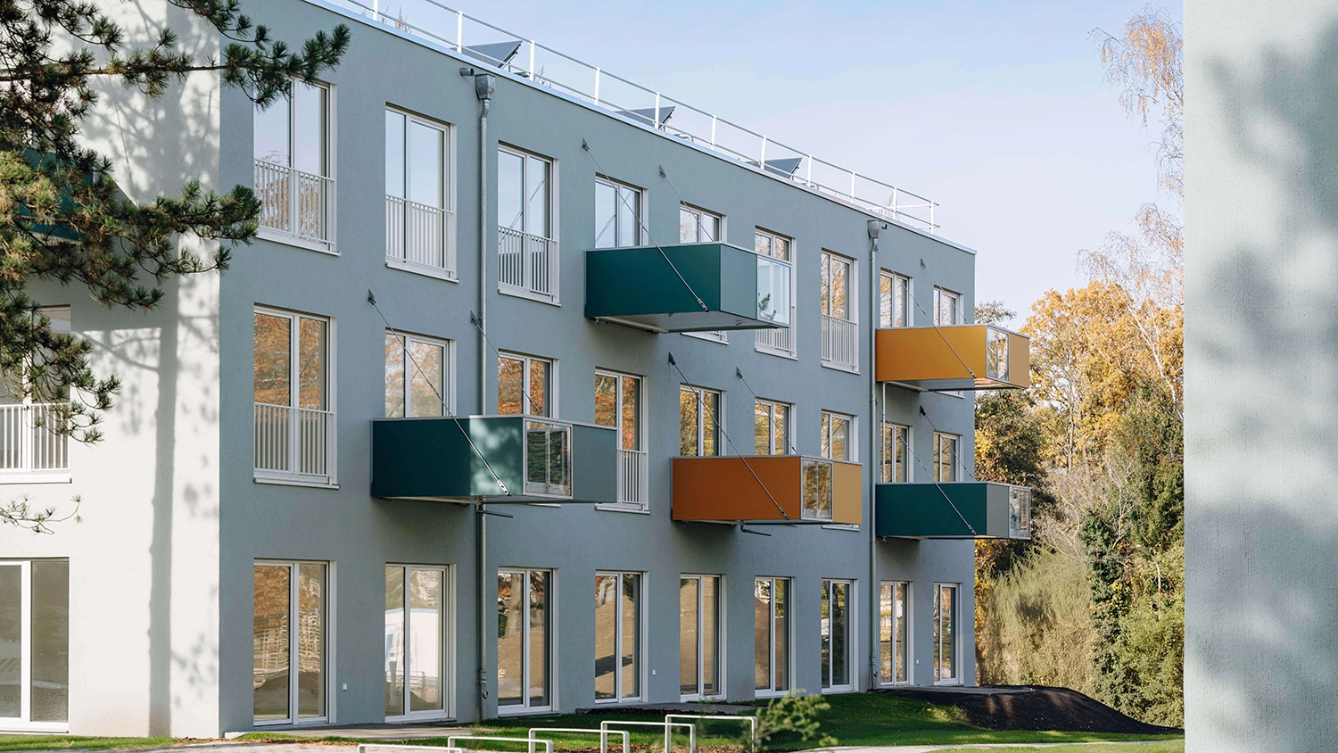 Modernes Wohngebäude mit grauer Fassade, farbigen Balkonen und Bäumen neben dem Gebäude