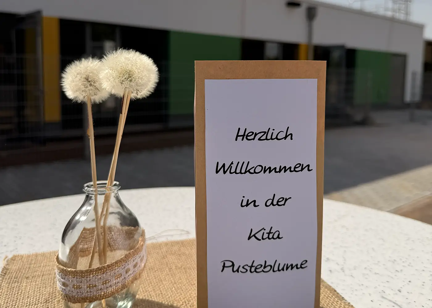 Dekorierte Tischszene vor einem Gebäude: Eine kleine Glasvase mit drei Pusteblumen steht neben einem Schild mit der Aufschrift ‚Herzlich Willkommen in der Kita Pusteblume‘