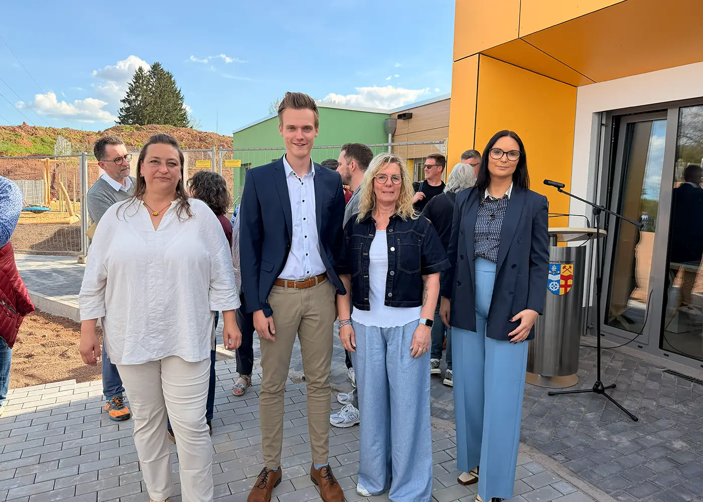 Vier Erwachsene stehen vor dem Eingang eines modernen Gebäudes bei einer Veranstaltung; im Hintergrund sind weitere Personen, ein Rednerpult mit Mikrofon und eine Baustelle zu sehen.